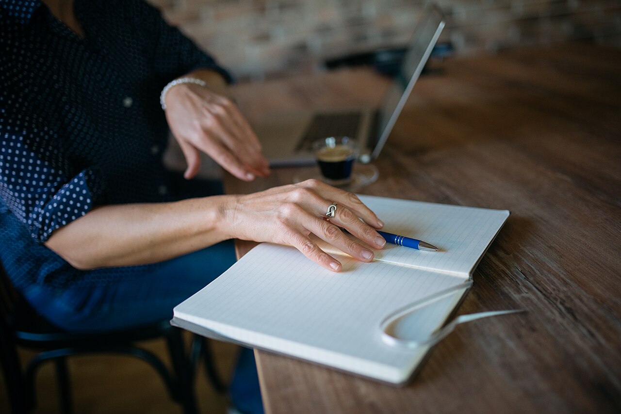 Close-up of a person placing a pen on a notebook. Laptop and a cup of espresso in a out of focus background. (c) Shixart1985, CC BY 2.0 <https://creativecommons.org/licenses/by/2.0>, via Wikimedia Commons