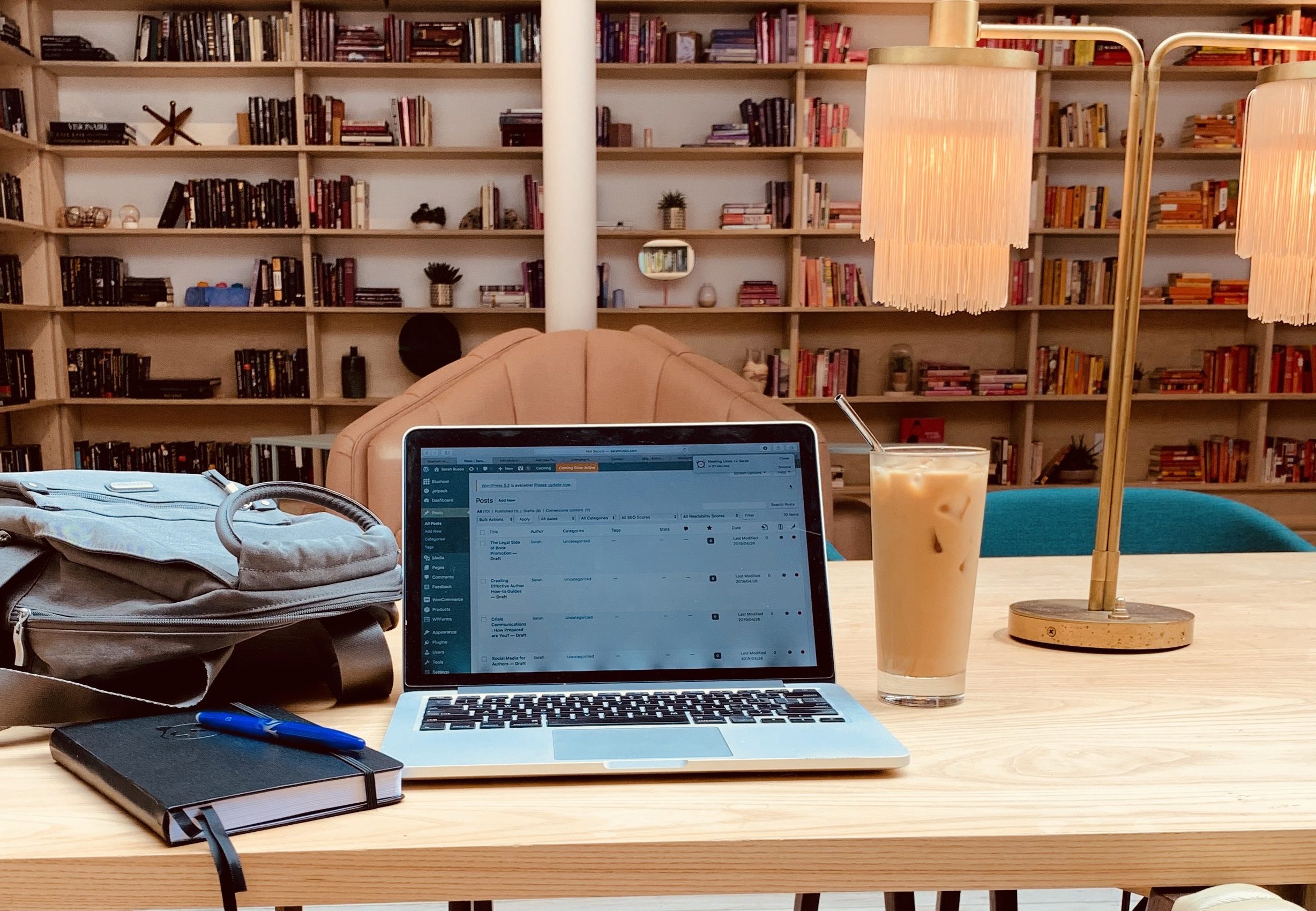 In the foreground a laptop, iced coffee, notebook and backpack with a lamp on a table. In the background, shelves covered with books and other objects. (wing)